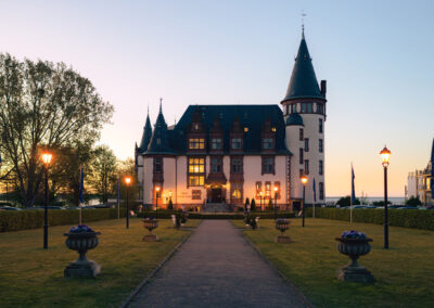 Schloss mit Türmen, gelegen an einem Seeufer mit Bäumen – romantische Außenansicht bei Abendlicht.