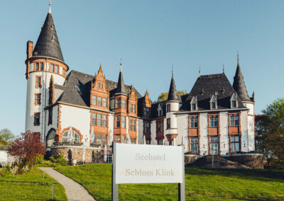 Frontaufnahme des Hotels mit Türmen und Rasen vor dem Eingang, dazu ein Schild mit dem Hotelnamen.